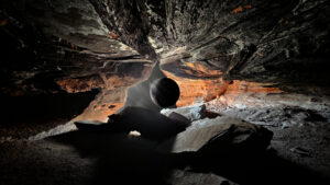 Inside a jagged cave, a large dark planet-like sphere hovers near the center against a rocky opening.