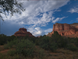Red sandstone cliffs and buttes rise above a desert shrub landscape under a partly cloudy blue sky.